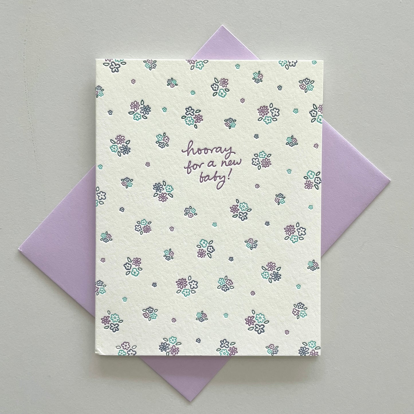 A floral baby letterpress greeting card with the text 'hooray for a new baby!' on it, displayed on a white surface with a purple envelope.