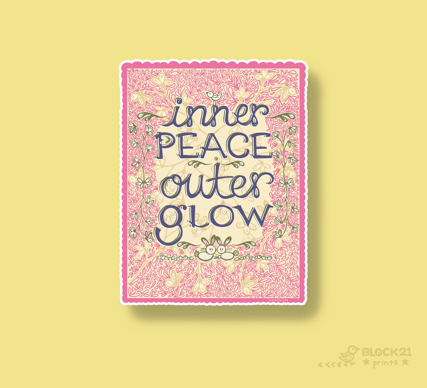 A square art print with a pink floral border and the text 'inner PEACE outer GLOW' in dark blue script, centered on a pale yellow background. A small 'BLOCK21' mark appears in the lower right corner.