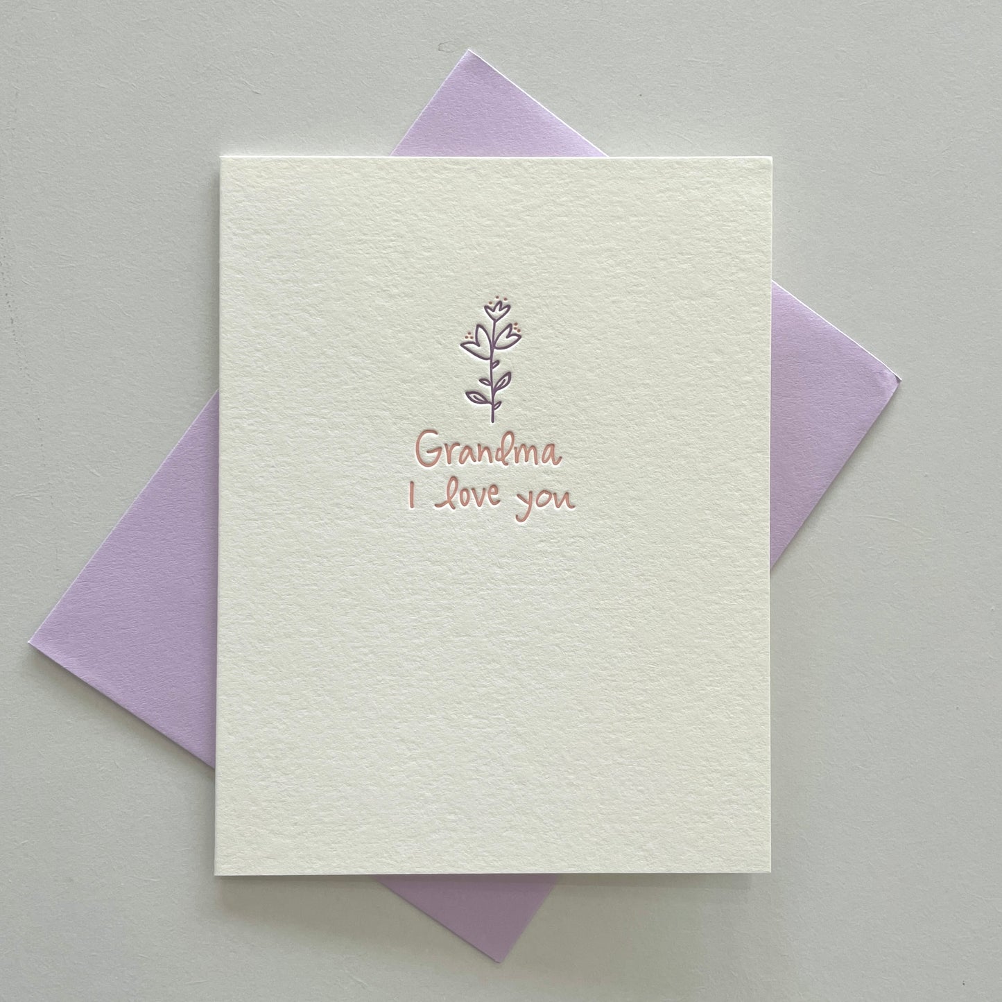 A white greeting card with the text 'Grandma I love you' printed in pink, accompanied by a purple envelope.