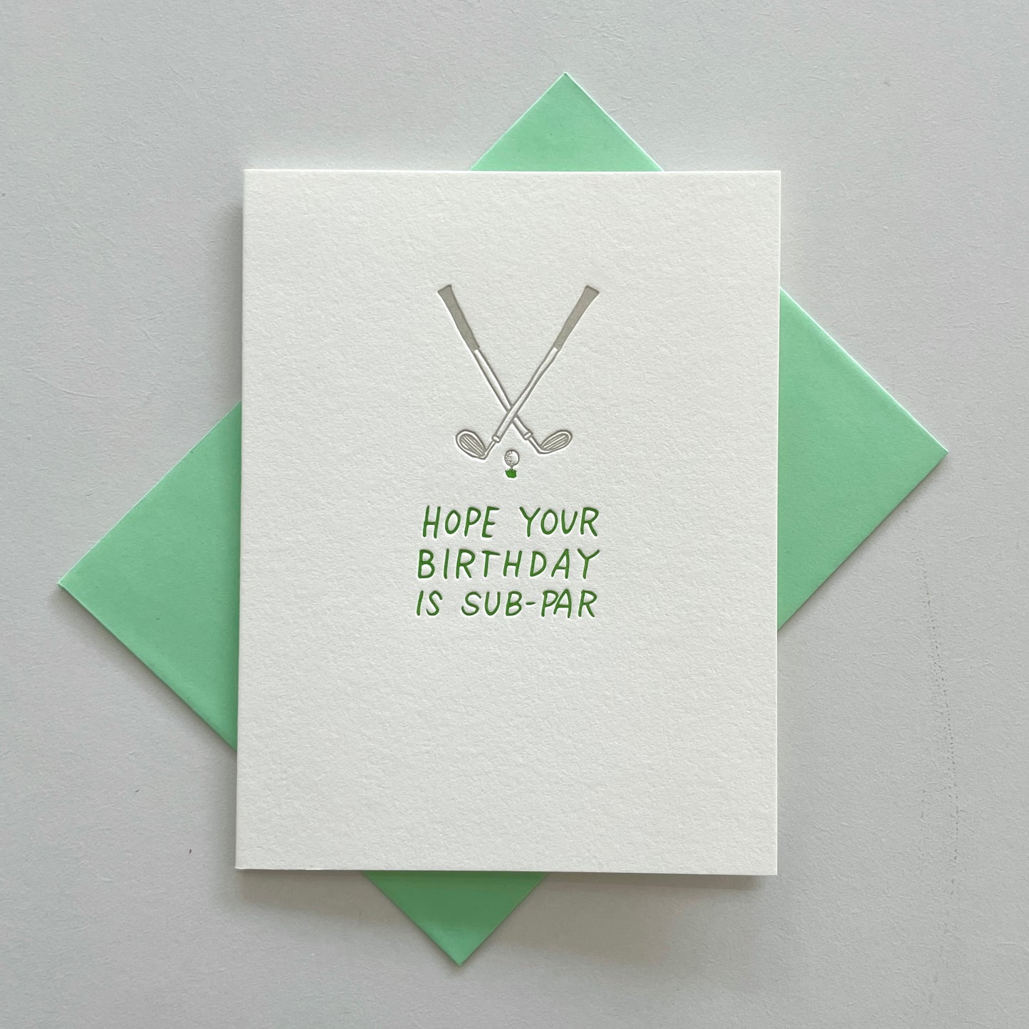 A letterpress greeting card with a golf-themed design and the text 'Hope your BIRTHDAY is SUB-PAR' on it. The card is placed on a green envelope against a grey background.