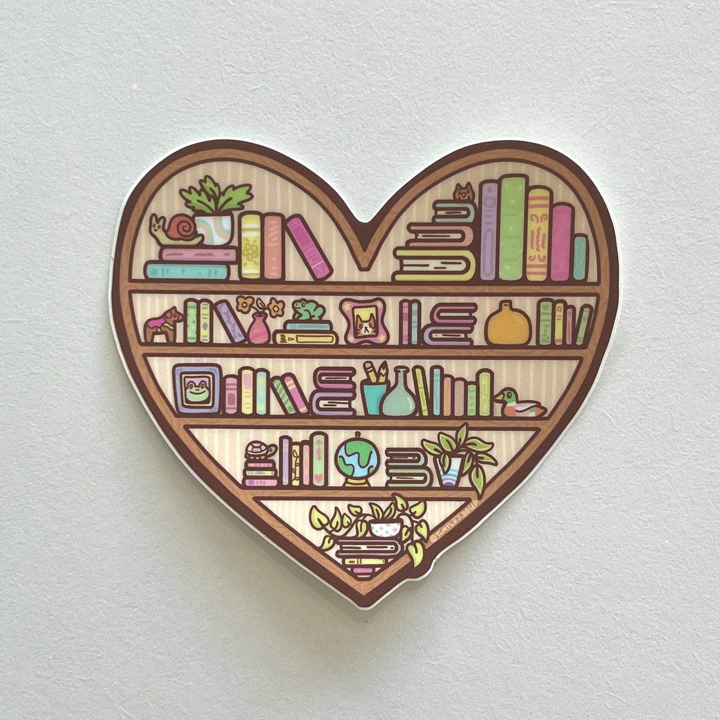 A heart-shaped vinyl sticker depicting a bookshelf with various books and decorative items.