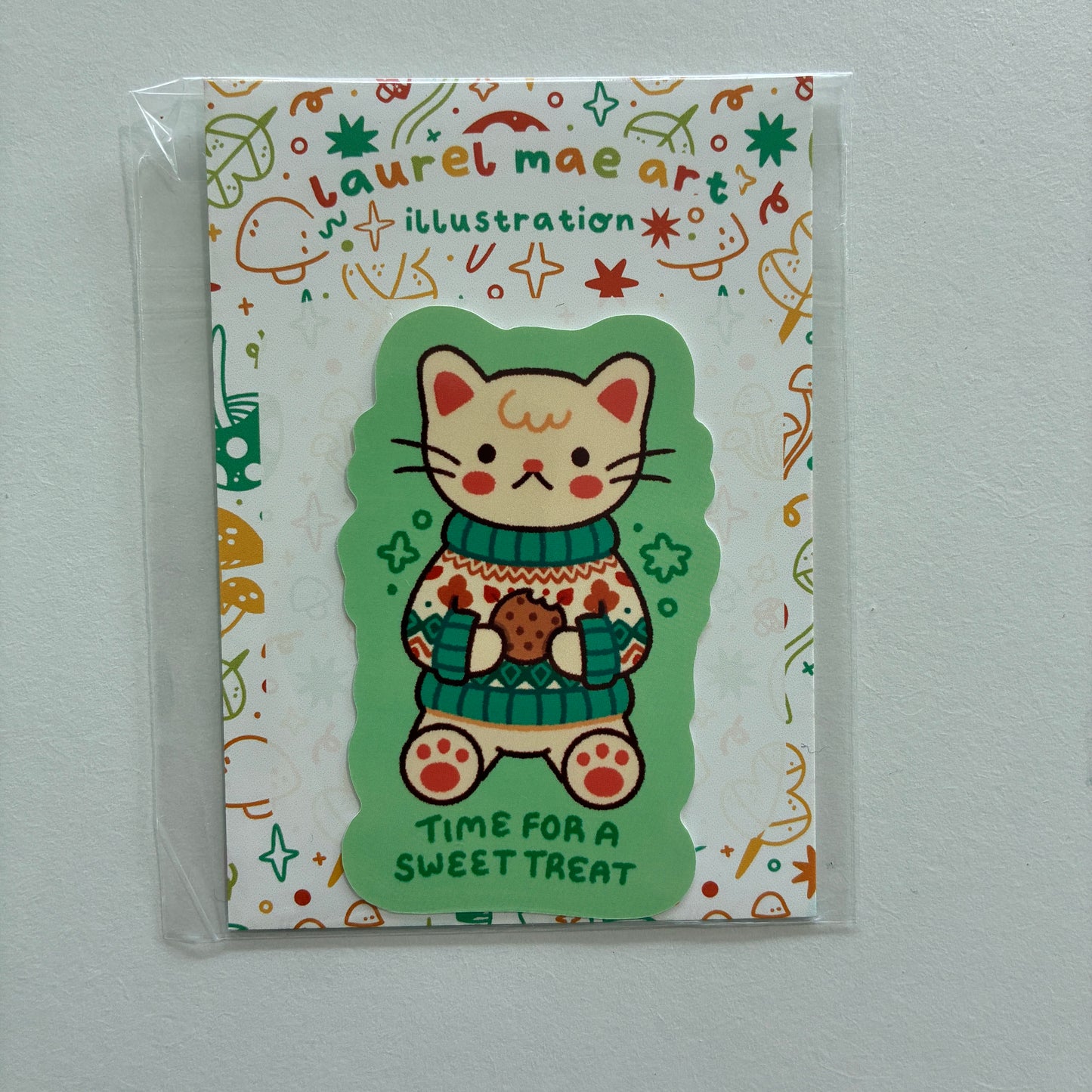 A packaged sticker featuring a cute cartoon cat in a green sweater holding a cookie, with the text 'TIME FOR A SWEET TREAT' on a white patterned backing card labeled 'Laurel Mae Art'.