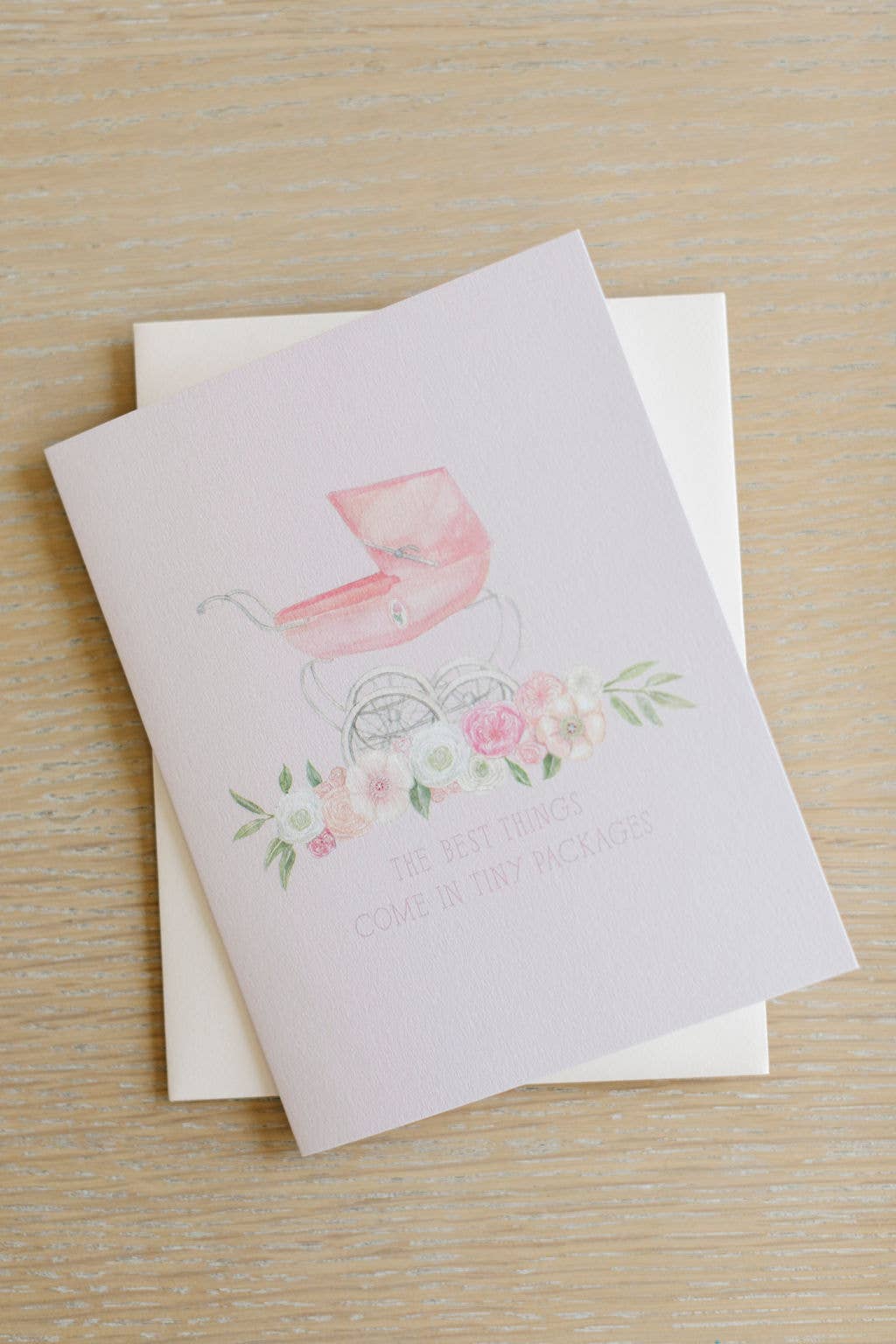 A baby shower card featuring a watercolor pink pram and floral design with the text 'THE BEST THINGS COME IN TINY PACKAGES', displayed on a light wood background with a white envelope behind it.