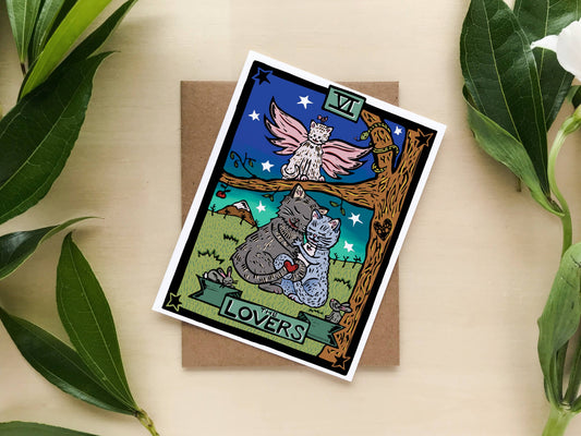 A tarot-style art print card titled 'LOVERS' with an illustrated gray cat and white cat under a tree, placed on a brown envelope and styled with green leaves on a light wood background.