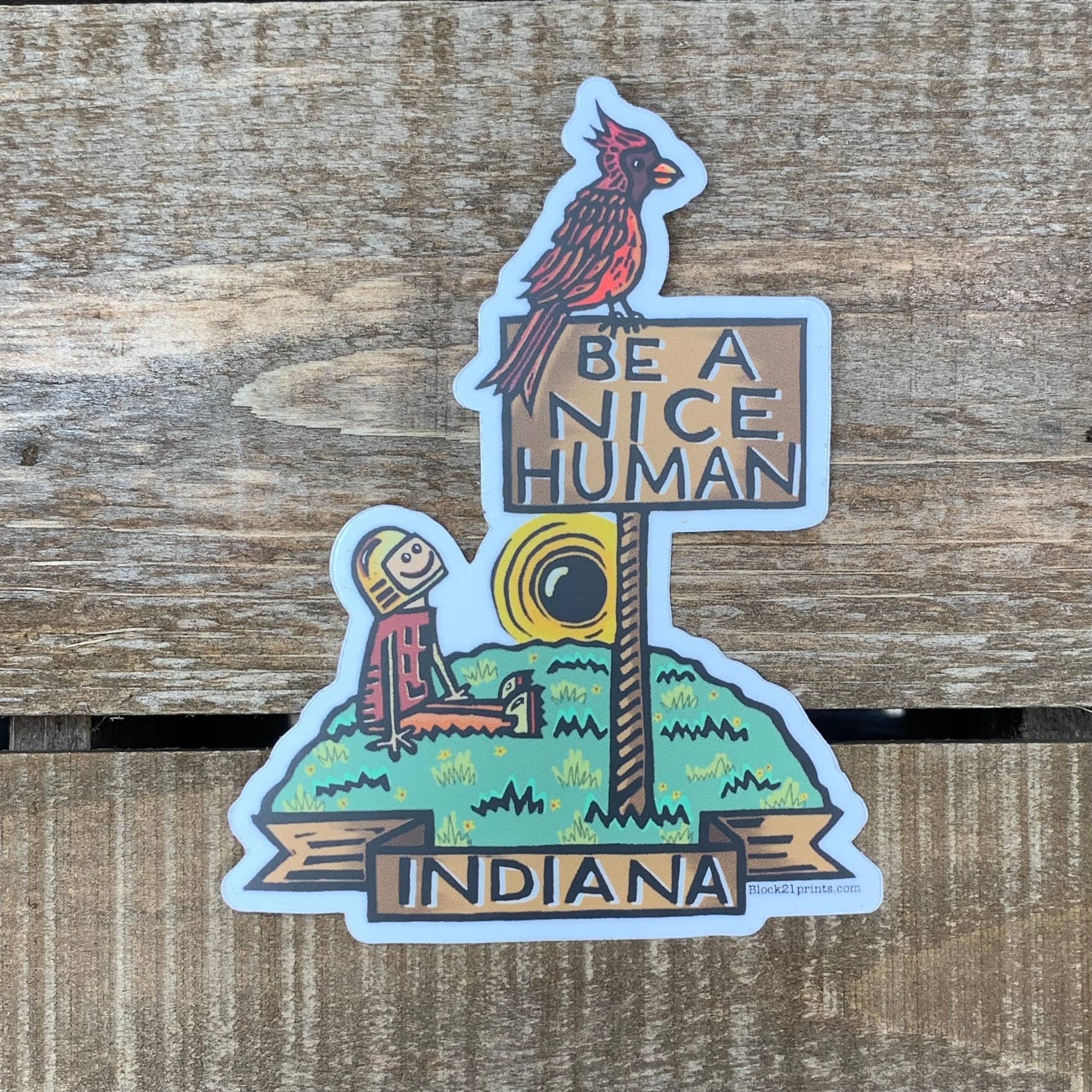 A die-cut sticker featuring a red cardinal perched on a sign that reads 'BE A NICE HUMAN' above a seated figure on a grassy hill, with 'INDIANA' on a banner below, placed on a rustic wooden surface.