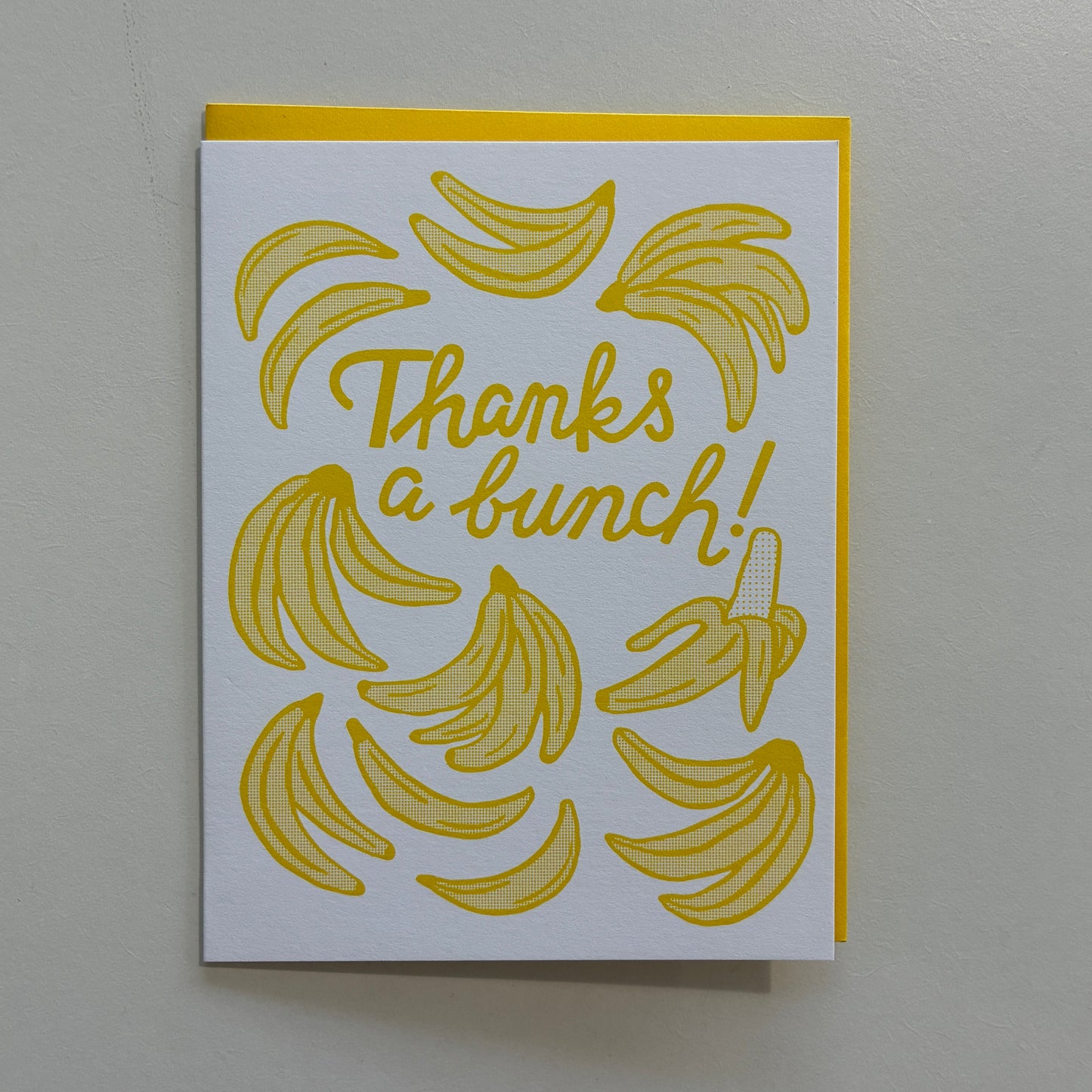 A white greeting card with a yellow border and yellow banana illustrations, featuring the text 'Thanks a bunch!' in yellow script, photographed against a light gray wall.
