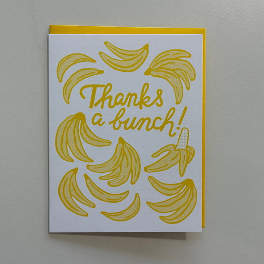 A white greeting card with a yellow border and yellow banana illustrations, featuring the text 'Thanks a bunch!' in yellow script, photographed against a light gray wall.
