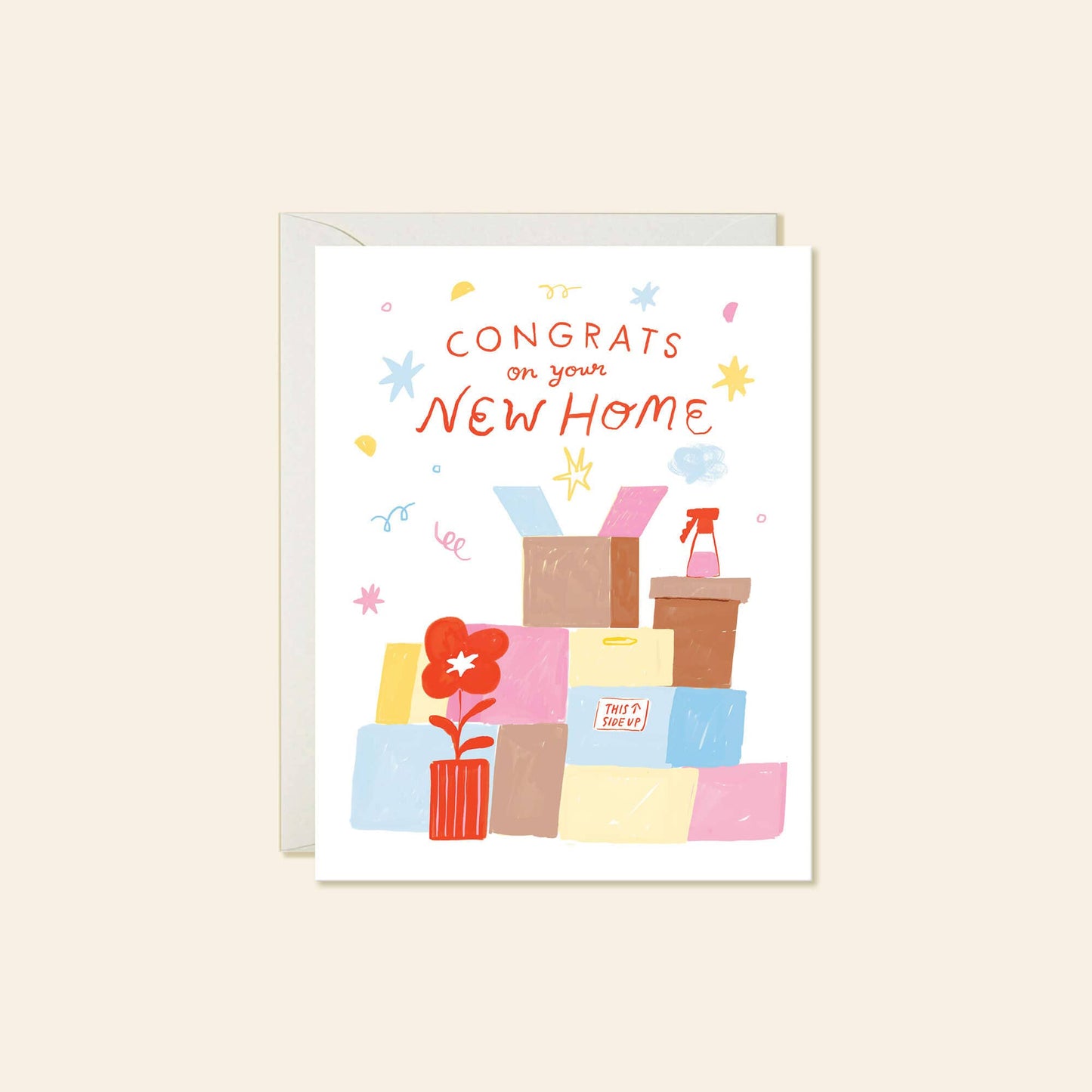 A white greeting card with a colorful illustration of moving boxes, a potted plant, and the text 'CONGRATS on your NEW HOME' in orange-red lettering, shown with a light gray envelope behind it on a warm beige background.