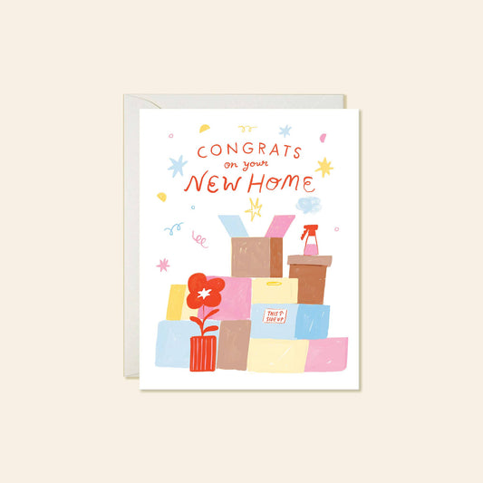 A white greeting card with a colorful illustration of moving boxes, a potted plant, and the text 'CONGRATS on your NEW HOME' in orange-red lettering, shown with a light gray envelope behind it on a warm beige background.
