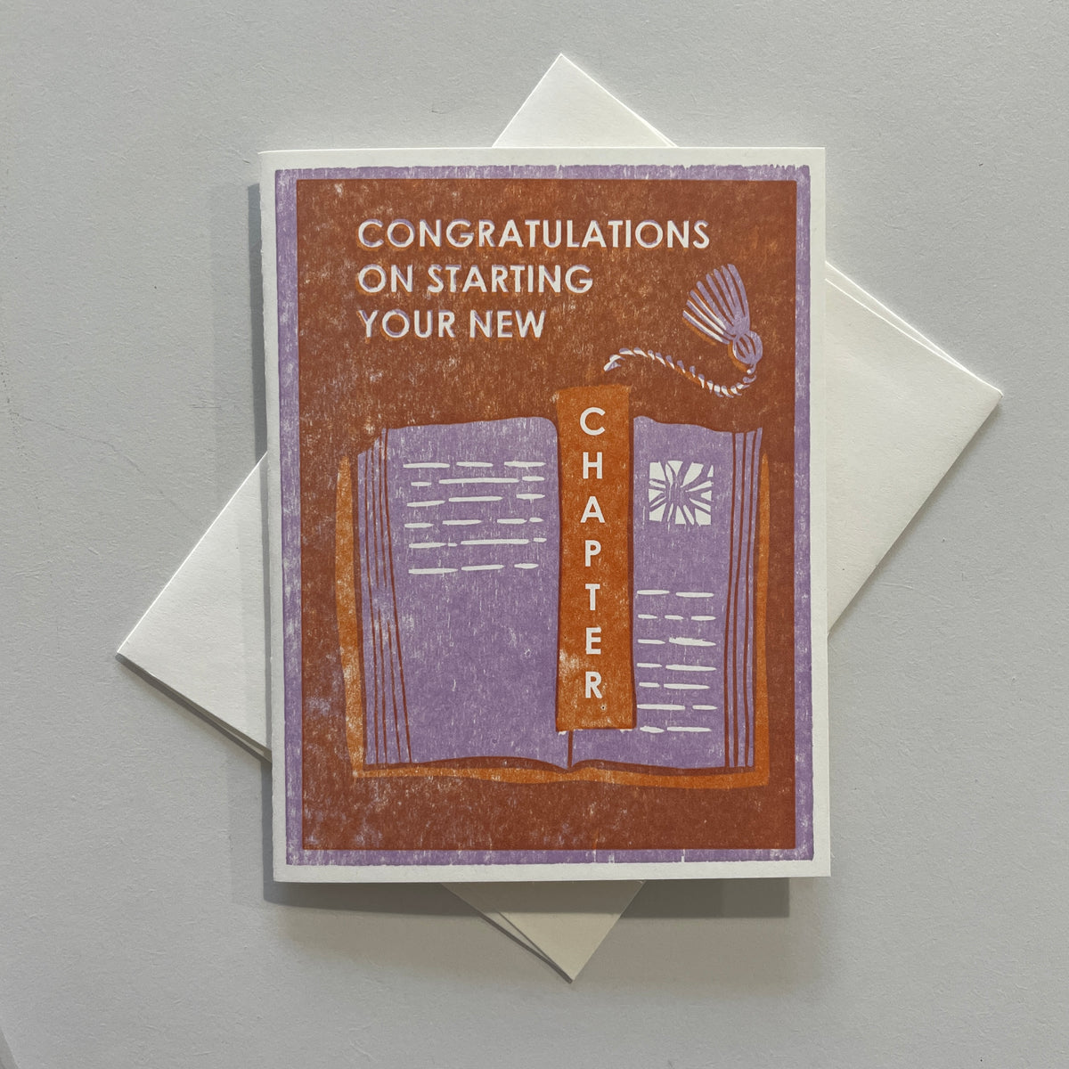 A greeting card with a letterpress printed message 'CONGRATULATIONS ON STARTING YOUR NEW CHAPTER' on a brown background, presented with an envelope.