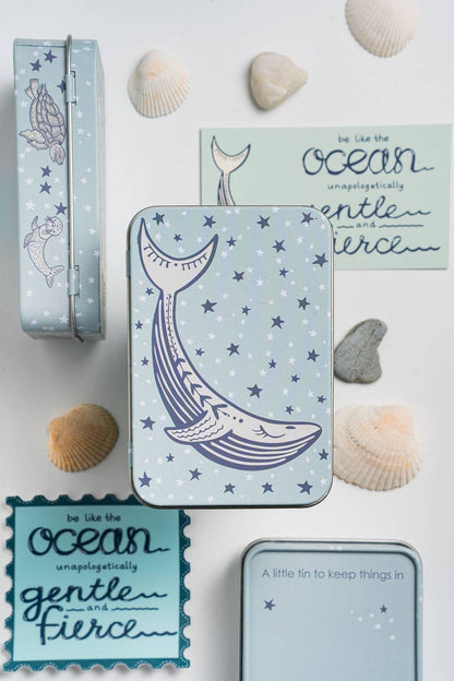 A styled flat-lay of a light blue metal tin with a whale illustration and star pattern, surrounded by seashells and matching decorative cards on a white background.