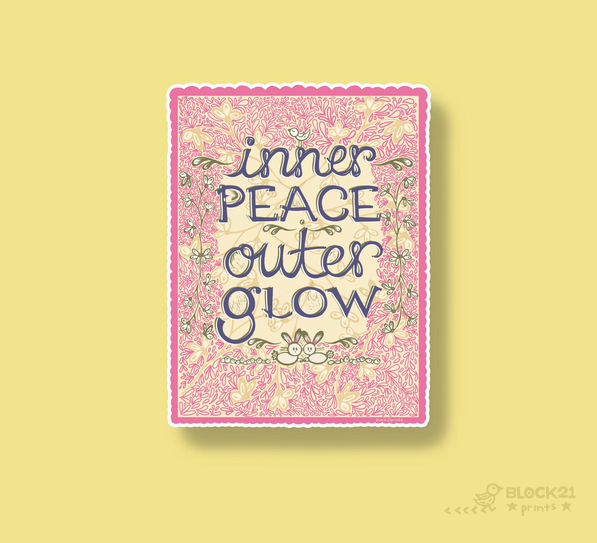 A square art print with a pink floral border and the text 'inner PEACE outer GLOW' in dark blue script, centered on a pale yellow background. A small 'BLOCK21' mark appears in the lower right corner.