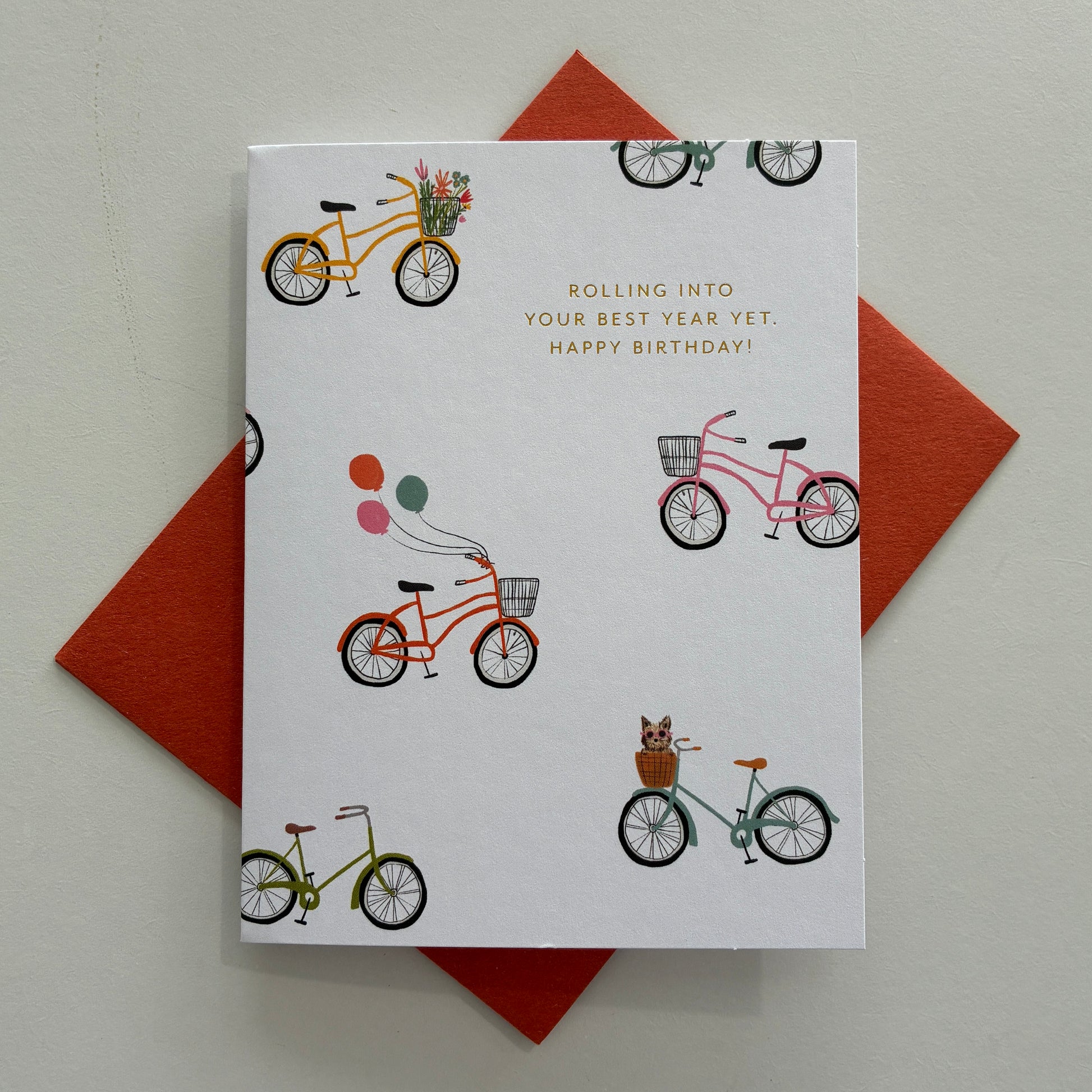 Greeting card with bicycle illustrations, gold foil text saying "Rolling into your best year yet. Happy Birthday!" and a red envelope on a light gray background