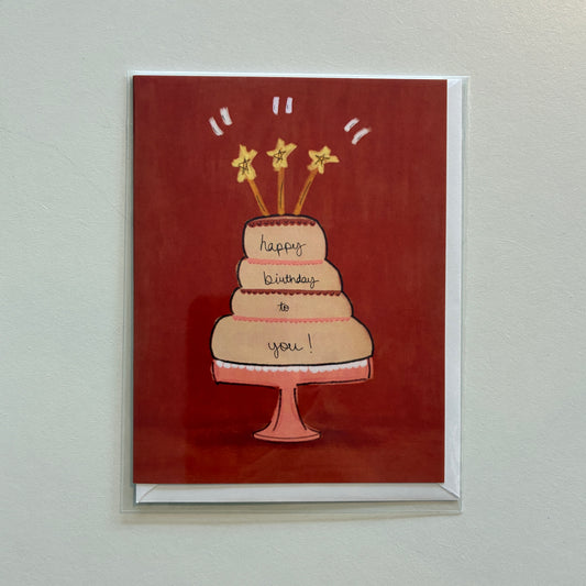 A square greeting card with a red background featuring an illustrated three-tier birthday cake on a stand, with candles and the words 'happy birthday to you!' written on the cake. The card is shown standing against a light neutral wall.