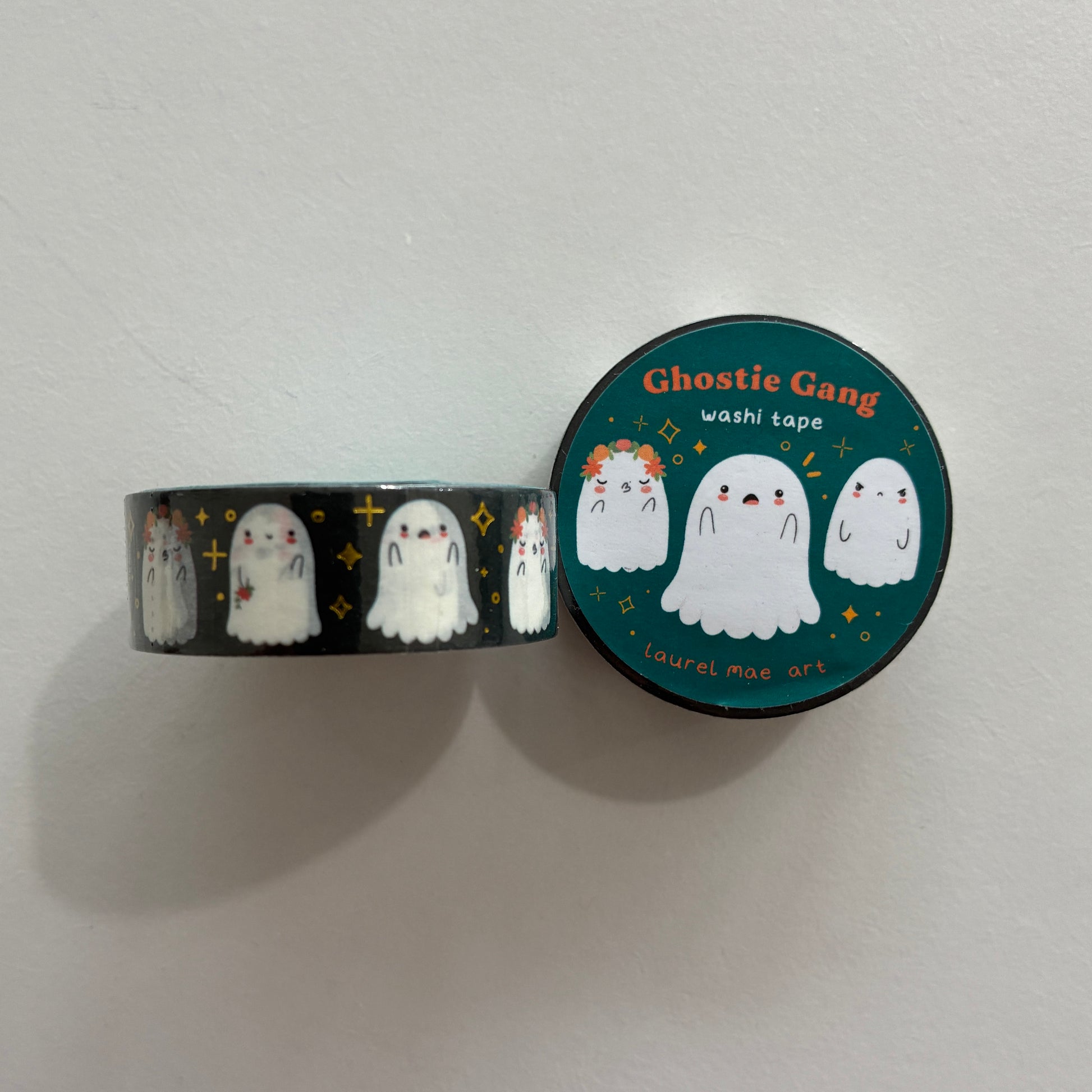 A roll of washi tape and its matching circular lid, featuring cute cartoon ghosts on a dark background. The lid reads 'Ghostie Gang washi tape' and shows three white ghost illustrations on a teal background with small yellow stars.