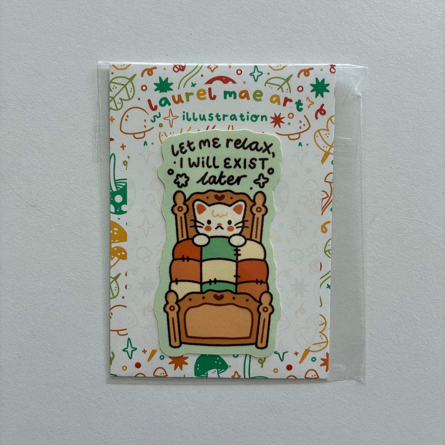 A cute die-cut sticker featuring a cartoon cat sitting in a wooden armchair and reading a green book, with the text 'Let me relax, I will exist later'. The sticker is displayed on its illustrated backing card by 'Laurel Mae Art', which is sealed in a clear plastic sleeve and photographed against a neutral light gray wall.