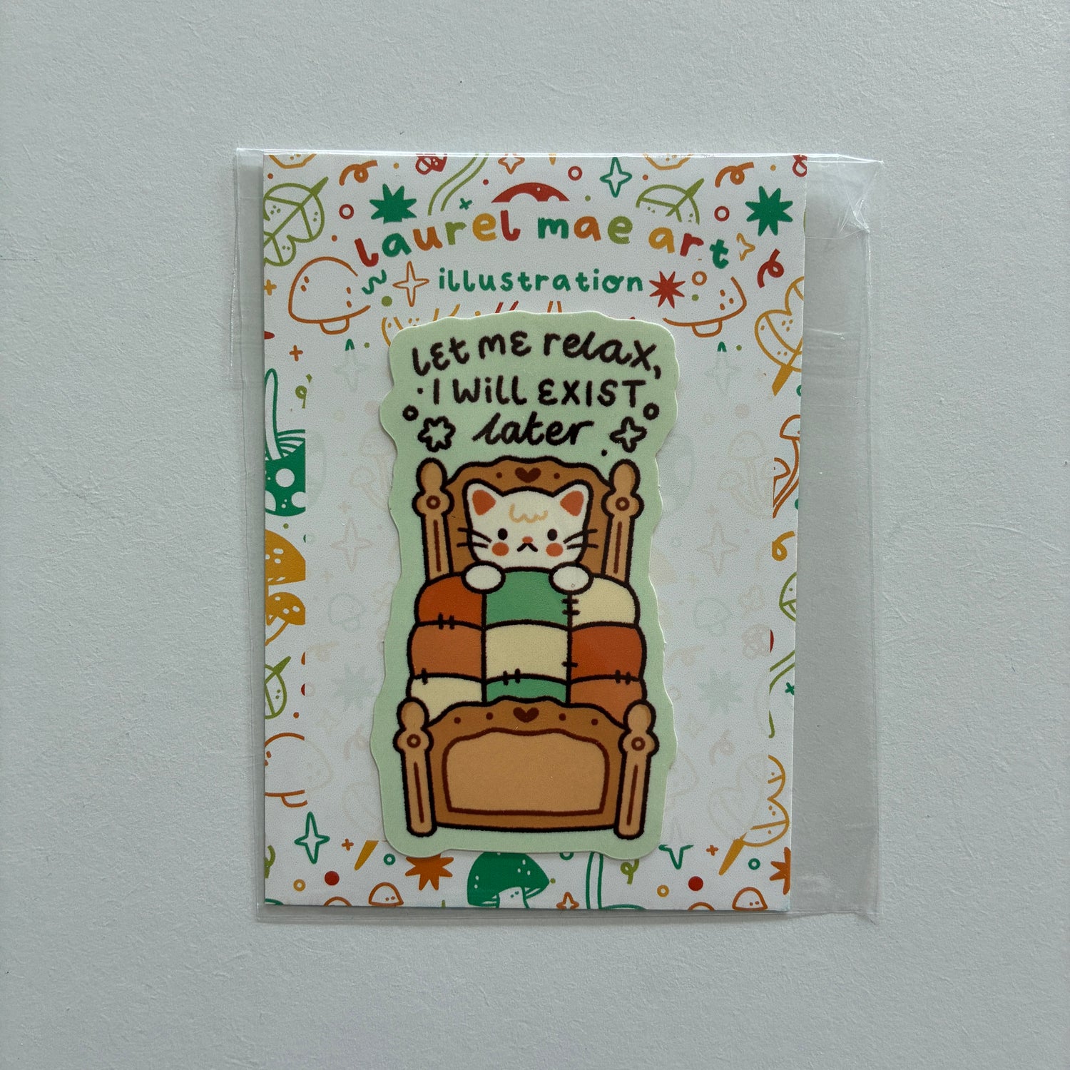 A cute die-cut sticker featuring a cartoon cat sitting in a wooden armchair and reading a green book, with the text 'Let me relax, I will exist later'. The sticker is displayed on its illustrated backing card by 'Laurel Mae Art', which is sealed in a clear plastic sleeve and photographed against a neutral light gray wall.