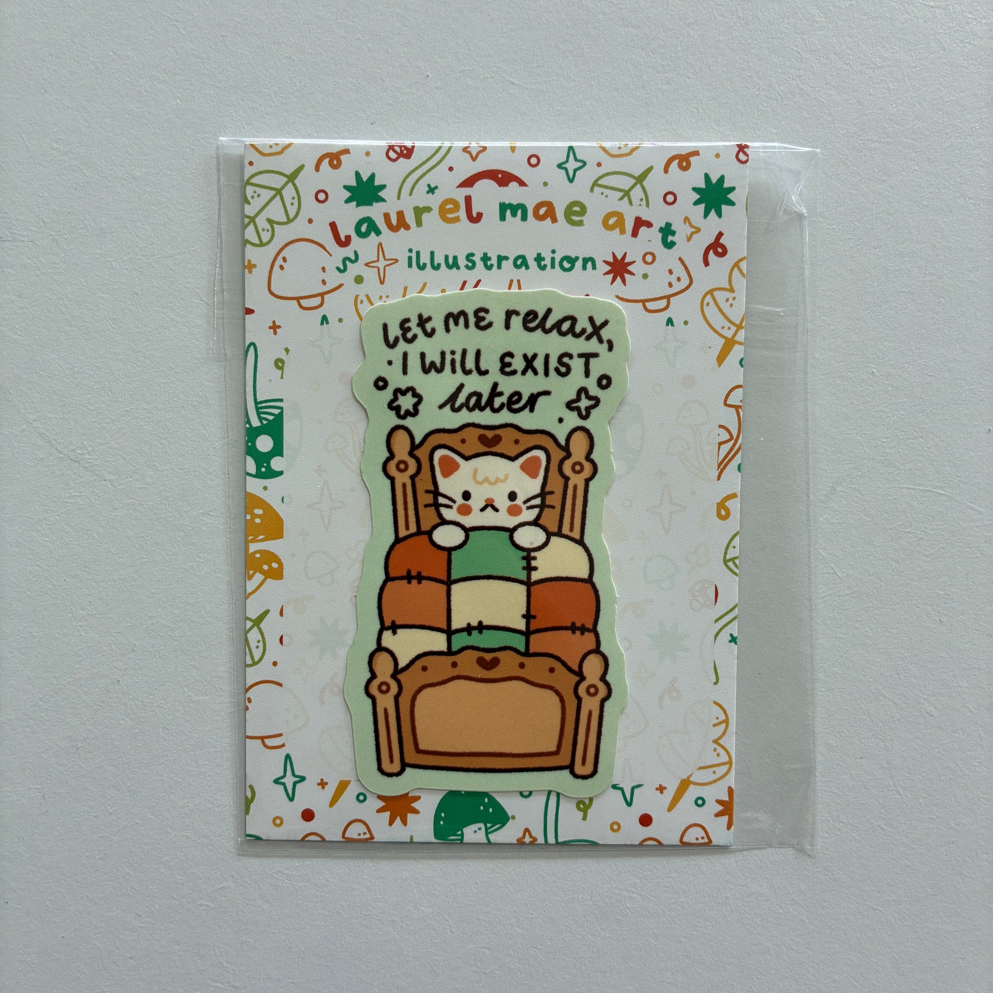 A cute die-cut sticker featuring a cartoon cat sitting in a wooden armchair and reading a green book, with the text 'Let me relax, I will exist later'. The sticker is displayed on its illustrated backing card by 'Laurel Mae Art', which is sealed in a clear plastic sleeve and photographed against a neutral light gray wall.