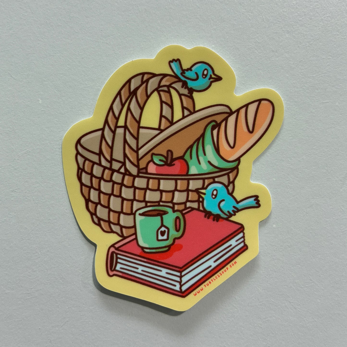 A sticker featuring a drawing of a picnic basket with a book on a gray background