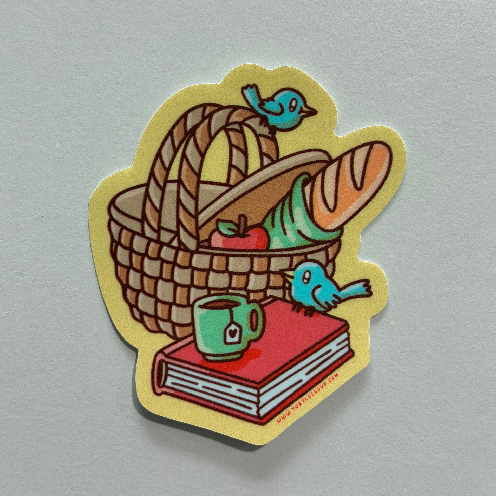 A sticker featuring a drawing of a picnic basket with a book on a gray background