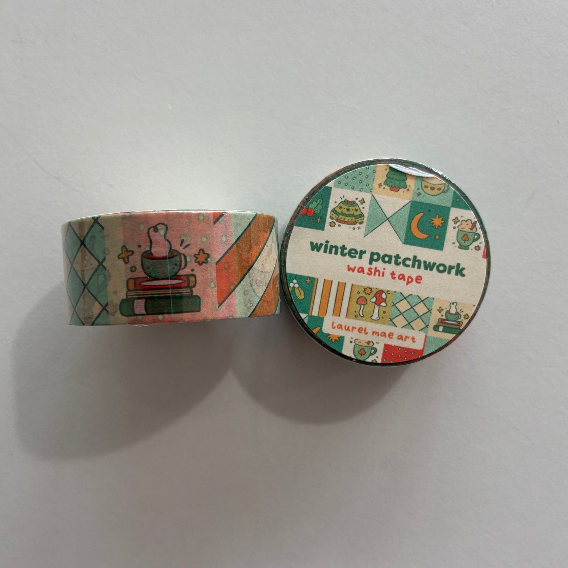 A roll and a round package of 'winter patchwork washi tape' by Laurel Mae Art, featuring a colorful winter-themed pattern with illustrations of a mug, snowman, and geometric shapes on a plain white background.