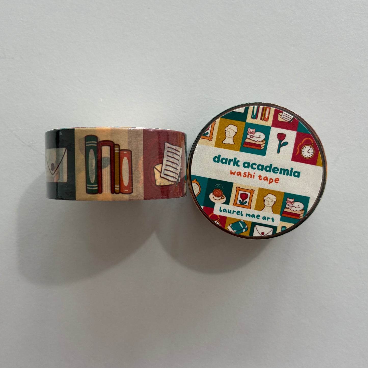 A roll of decorative washi tape with a dark academia theme, shown partially unrolled next to its round lid. The pattern features books, notebooks, coffee cups, and flowers in muted red, green, beige, and brown tones.