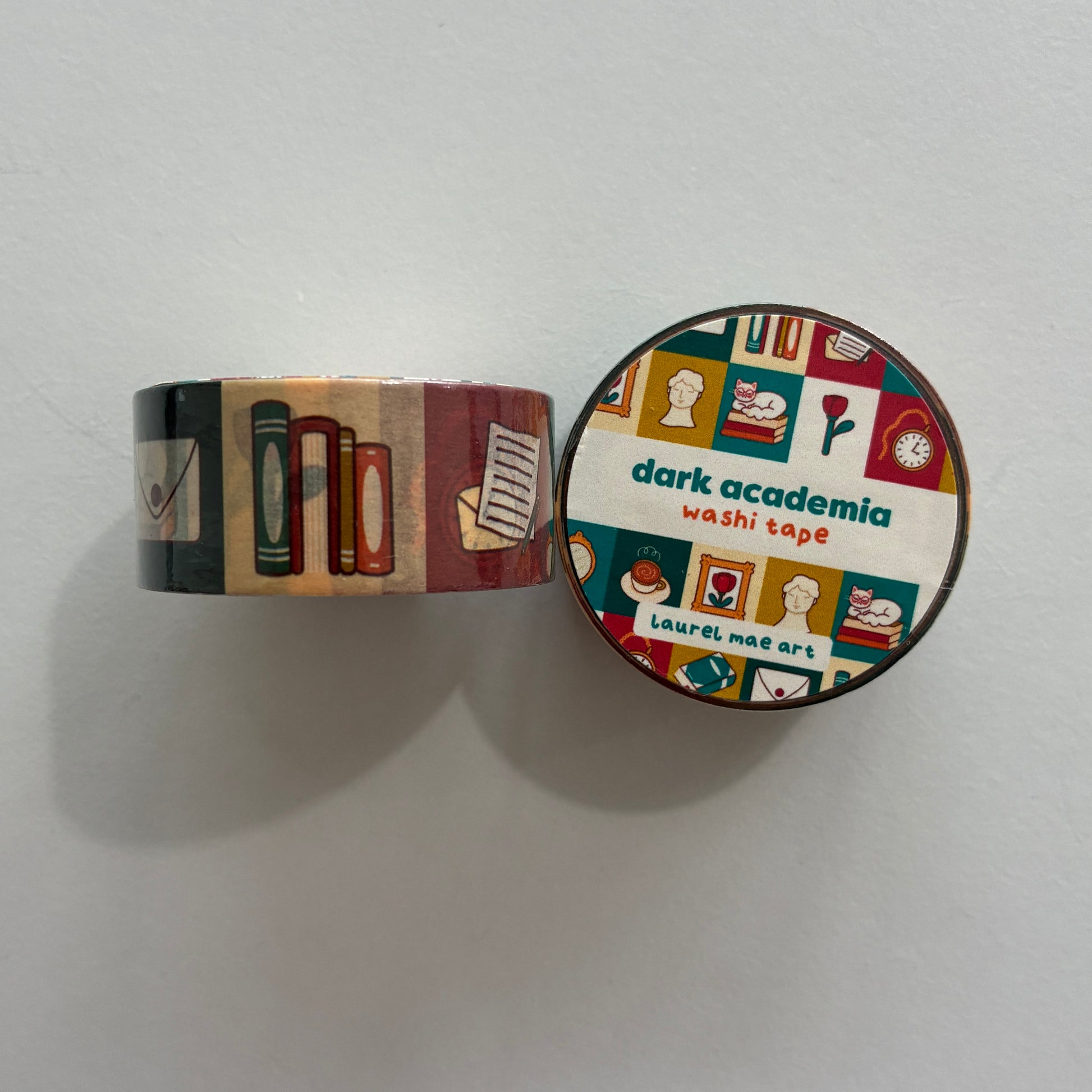A roll of decorative washi tape with a dark academia theme, shown partially unrolled next to its round lid. The pattern features books, notebooks, coffee cups, and flowers in muted red, green, beige, and brown tones.