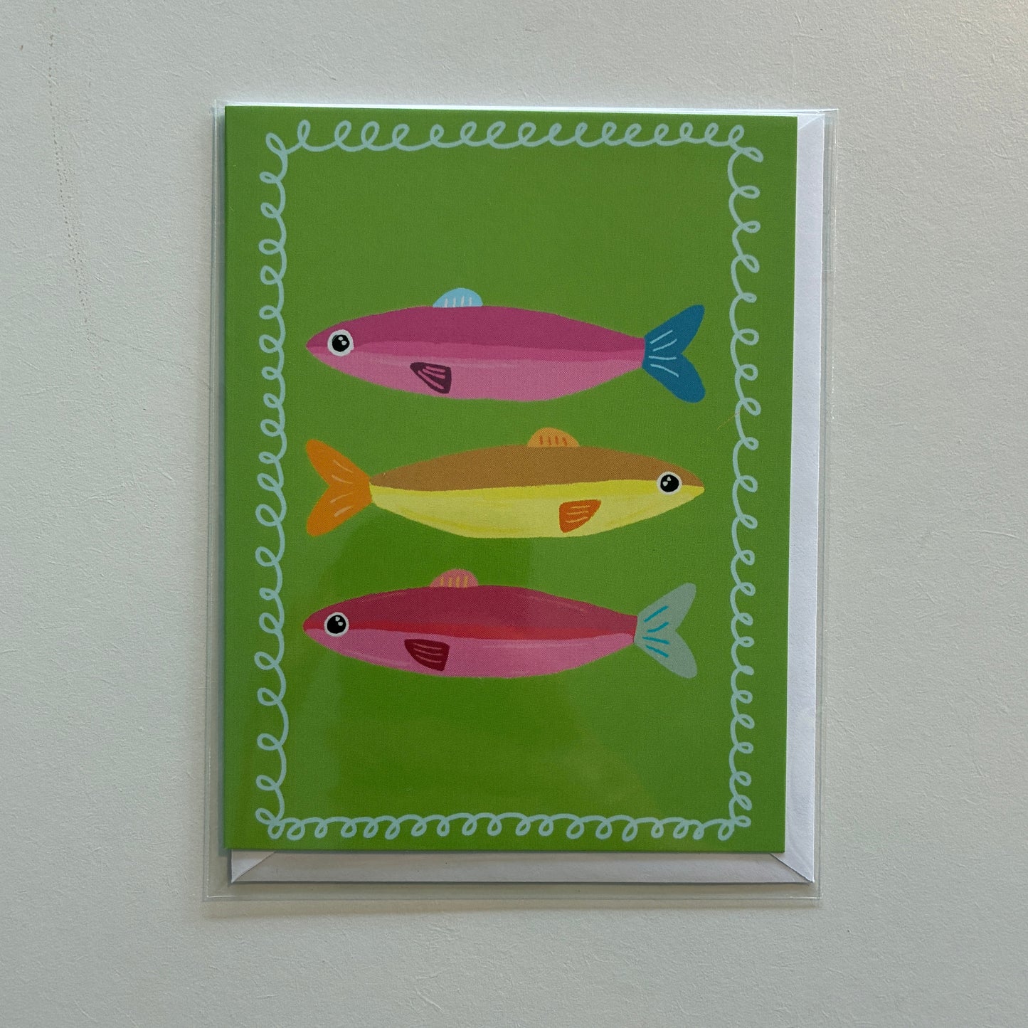 A square greeting card featuring three stylized fish in pink, yellow, and orange on a bright green background, bordered by a white wavy pattern. The card is shown hanging on a light gray wall.