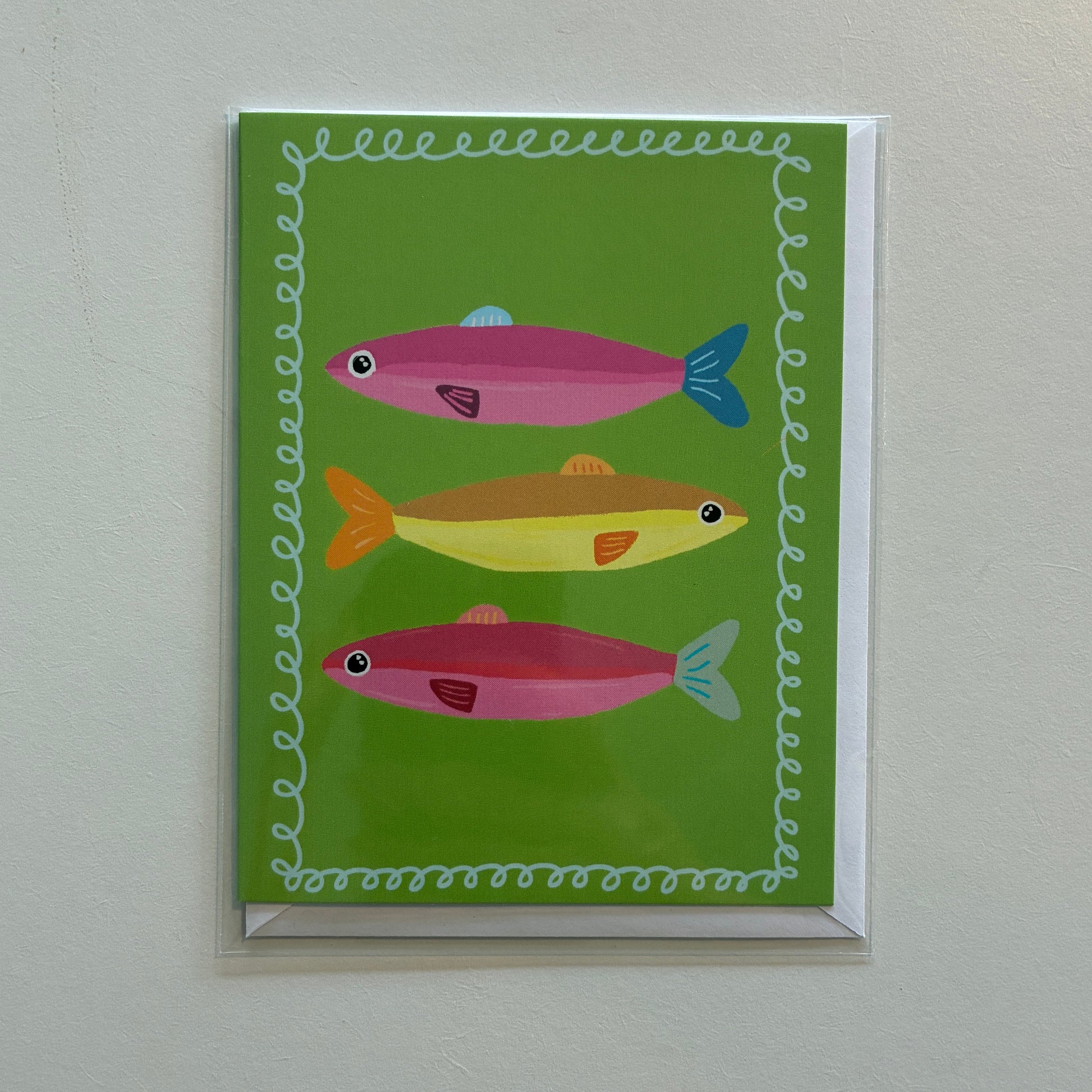 A square greeting card featuring three stylized fish in pink, yellow, and orange on a bright green background, bordered by a white wavy pattern. The card is shown hanging on a light gray wall.