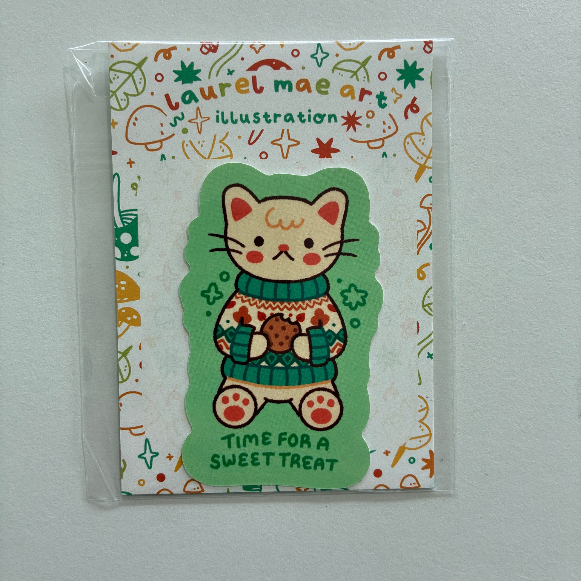 A packaged sticker featuring a cute cartoon cat in a green sweater holding a cookie, with the text 'TIME FOR A SWEET TREAT' on a white patterned backing card labeled 'Laurel Mae Art'.
