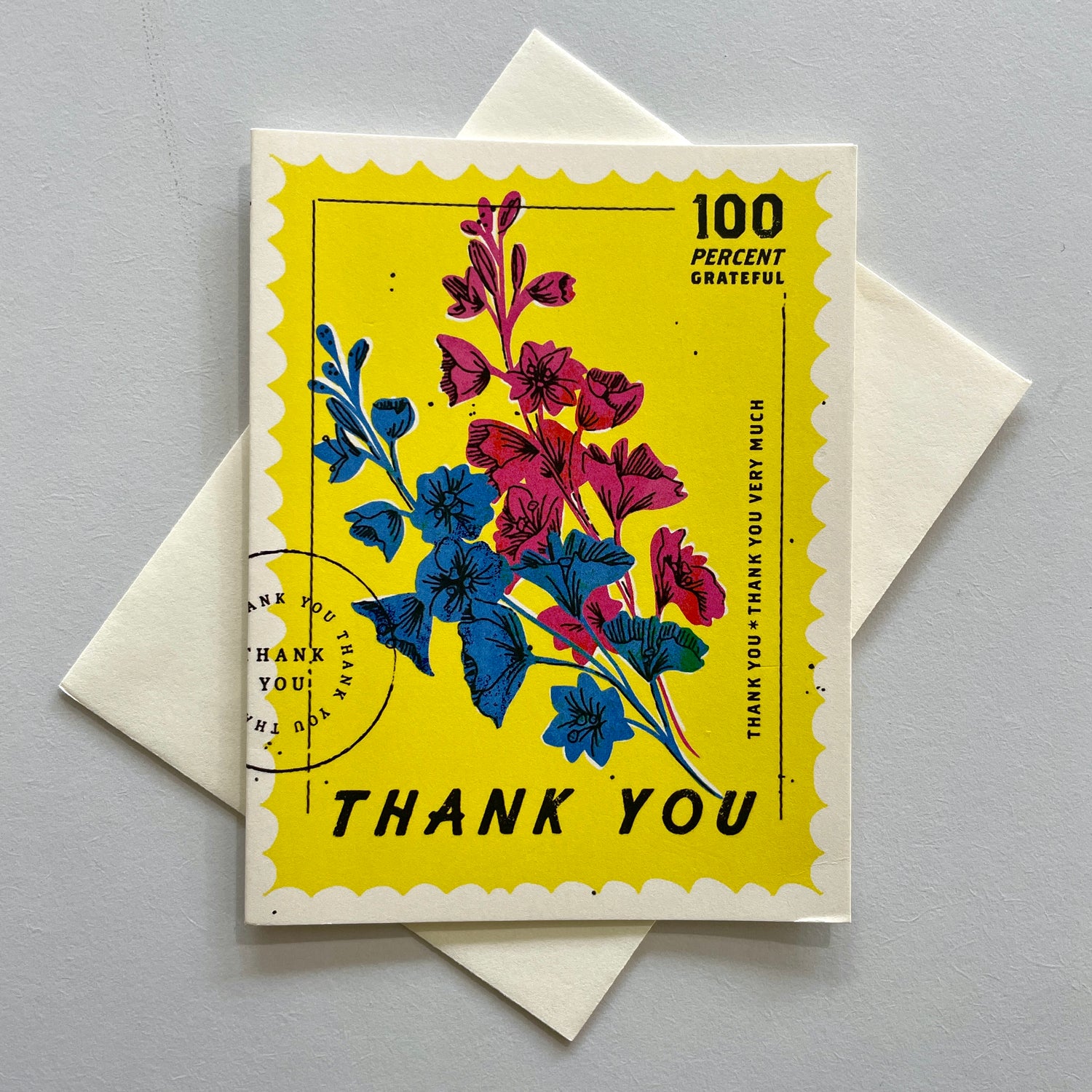 A greeting card with a colorful floral design and the text 'THANK YOU' on it, placed on a white surface with a postage stamp and a white envelope.