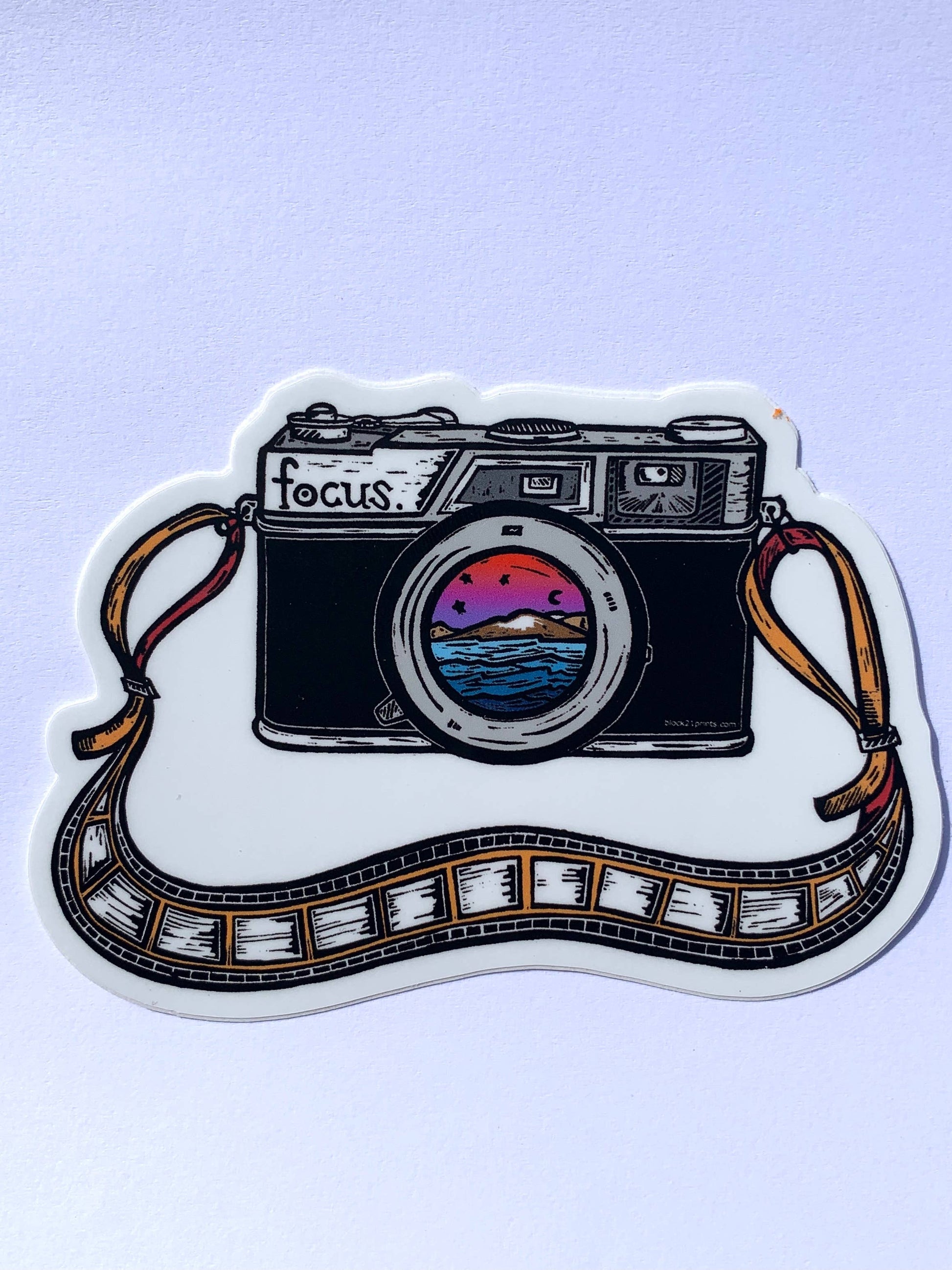A die-cut sticker featuring a hand-drawn camera with a colorful sunset reflection in its lens, a film strip wrapping around the bottom, and the word 'focus.' on the camera body, shown on a light lavender background.