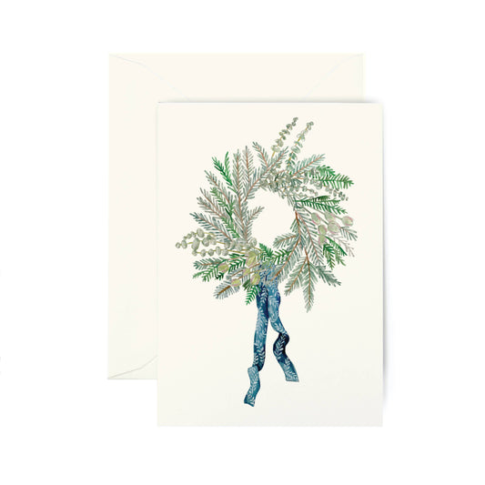 A cream-colored greeting card featuring an illustration of a winter wreath with green and silver pine branches tied with a blue ribbon, shown with a matching cream envelope behind it on a white background.