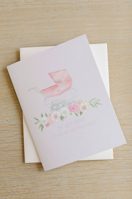 A baby shower card featuring a watercolor pink pram and floral design with the text 'THE BEST THINGS COME IN TINY PACKAGES', displayed on a light wood background with a white envelope behind it.