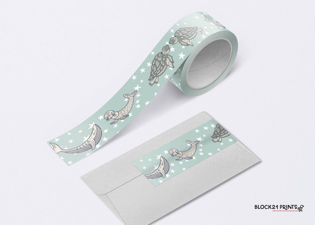 A roll of washi tape with a sea life pattern, shown partially unrolled next to a light gray envelope. The tape features illustrations of dolphins, seals, and turtles on a pale mint green background with small white stars.